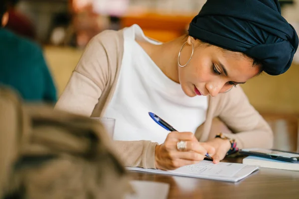 young, attractive Muslim student (Arab, Malay, Asian) writing in notepad in cafe in the day. She is wearing a turban (headscarf, hijabi) and is elegantly dressed in earthy tones.