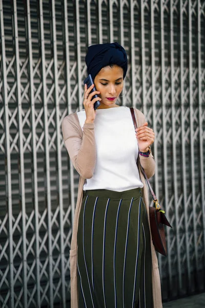 Portrait of a smartly dressed Muslim (Asian, Malay, Arab) woman wearing a turban (hijab, headscarf) with smartphone. She is smartly dressed in a camisole and cardigan against a metal door.