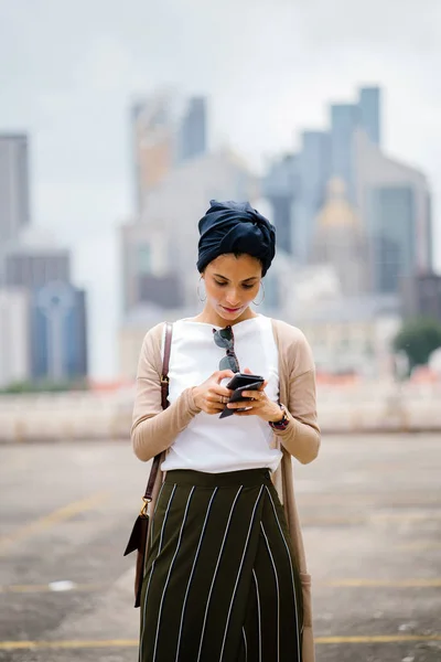 アラブ マレー語 アジア 若いイスラム教徒の女性の肖像画の背景 シンガポール市街のスカイラインに対してスマート フォン ヒジャーブ ヘッド スカーフ ターバンを着て — ストック写真