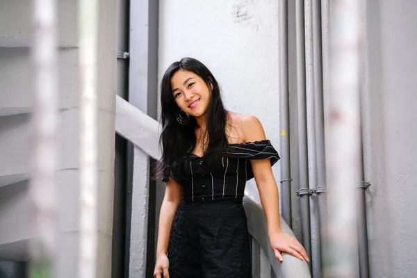 Portrait of a young Chinese woman lawyer (Singaporean) standing on vintage white steps in her office or coworking space. She is professionally dressed in a black outfit and is smiling at the camera.