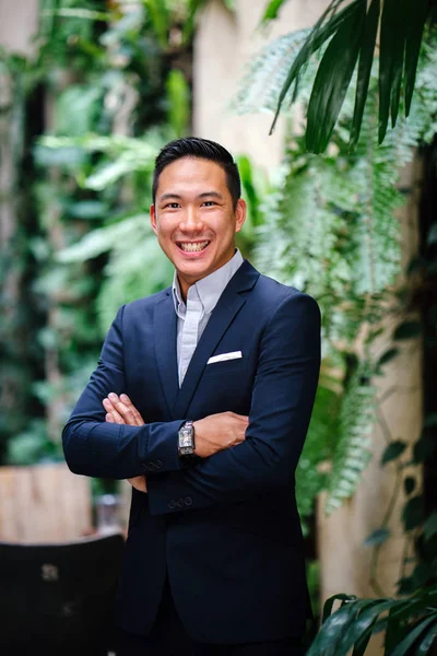 Portrait of a handsome, young, smart Chinese Asian man in a suit smiling and standing against a green wall in the day. He has short hair and is wearing a grey suit.