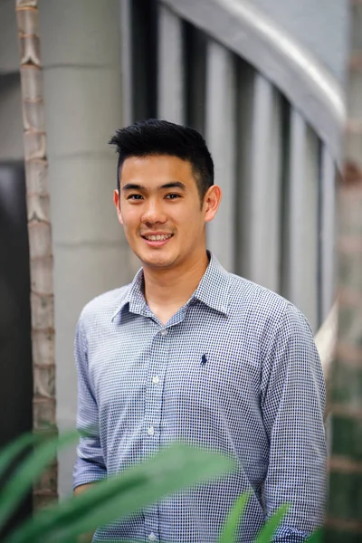 Portrait of a handsome, young, smart Chinese Asian man in a suit smiling and standing against a green wall in the day. He has short hair and is wearing a grey suit.