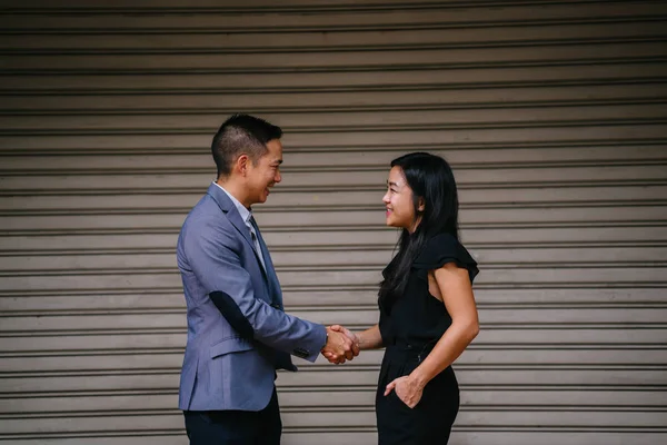 Two Chinese business people (a man in a suit and professionally dressed woman) shake hands in agreement and accord over a deal in the daytime against a plain background.