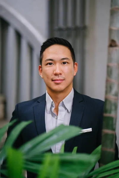 Portrait of a handsome, young, smart Chinese Asian man in a suit smiling and standing against a green wall in the day. He has short hair and is wearing a grey suit.