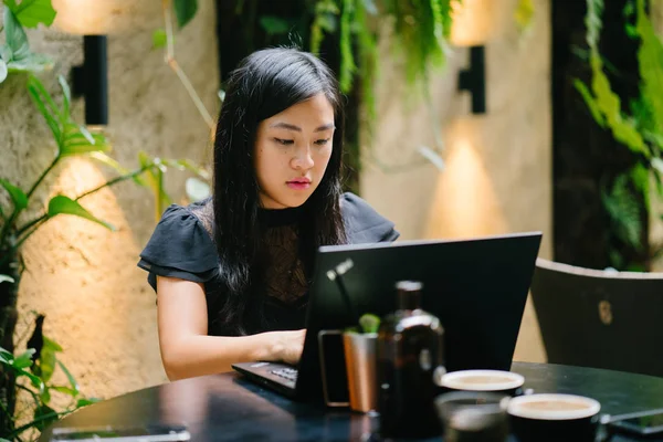 A young Chinese woman lawyer is working on her laptop in her office, coworking space or her office with some coffee and drinks on a table. She is smiling as she focuses on her work.