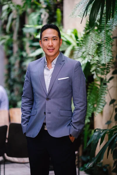 Portrait of a handsome, young, smart Chinese Asian man in a suit smiling and standing against a green wall in the day. He has short hair and is wearing a grey suit.