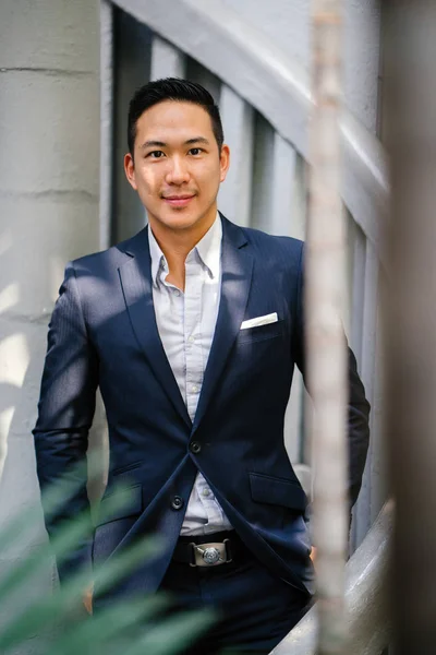 Portrait of a handsome, young, smart Chinese Asian man in a suit smiling and standing against a green wall in the day. He has short hair and is wearing a grey suit.