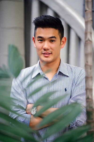 Portrait of a handsome, young, smart Chinese Asian man in a suit smiling and standing against a green wall in the day. He has short hair and is wearing a grey suit.