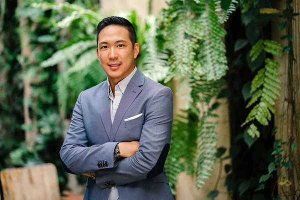 Portrait of a handsome, young, smart Chinese Asian man in a suit smiling and standing against a green wall in the day. He has short hair and is wearing a grey suit.