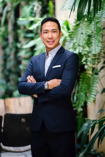 Portrait of a handsome, young, smart Chinese Asian man in a suit smiling and standing against a green wall in the day. He has short hair and is wearing a grey suit.