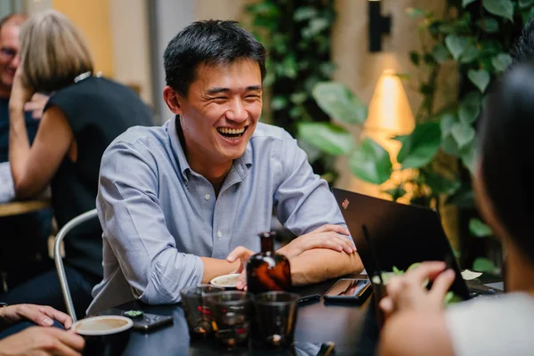 group of young and energetic Chinese Asian coworkers sit around a table and have a light hearted business discussion. They are smiling and laughing as they talk and have a conversation.