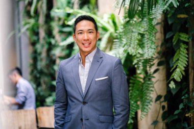 Yakışıklı, genç, akıllı portresi Çinli Asyalı adam gülümseyerek ve gün yeşil bir duvara karşı duran bir takım. Kısa saçlı ve gri takım elbise giyiyor.