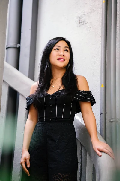 Portrait of a young Chinese woman lawyer (Singaporean) standing on vintage white steps in her office or coworking space. She is professionally dressed in a black outfit and is smiling at the camera.