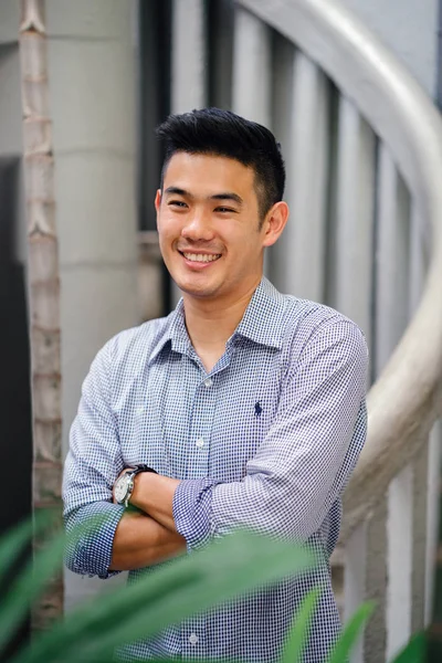 Portrait of a handsome, young, smart Chinese Asian man in a suit smiling and standing against a green wall in the day. He has short hair and is wearing a grey suit.