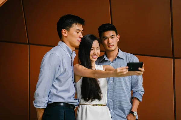 group of Asian coworkers pose and take a sefie / selfie photograph in the day against a futuristic backdrop. They are smiling, relaxed, young and attractive. Two men and a woman pose for the photo.