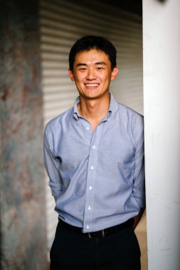 Yakışıklı, genç, akıllı portresi Çinli Asyalı adam gülümseyerek ve gün duvara karşı duran bir takım. Kısa saçlı ve gri takım elbise giyiyor.