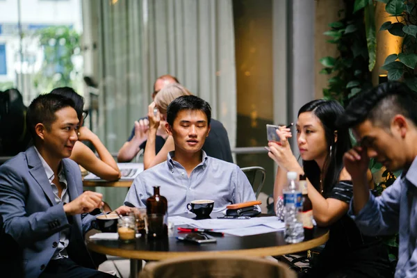 group of young and energetic Chinese Asian coworkers sit around a table and have a light hearted business discussion. They are smiling and laughing as they talk and have a conversation.