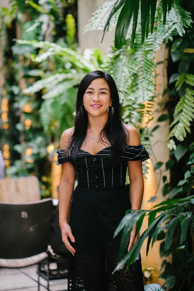 Portrait of a young Chinese woman lawyer (Singaporean) standing near a wall in her office with plants and leaves. She is professionally dressed and is smiling at the camera.