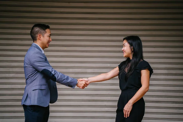 Two Chinese business people (a man in a suit and professionally dressed woman) shake hands in agreement and accord over a deal in the daytime against a plain background.