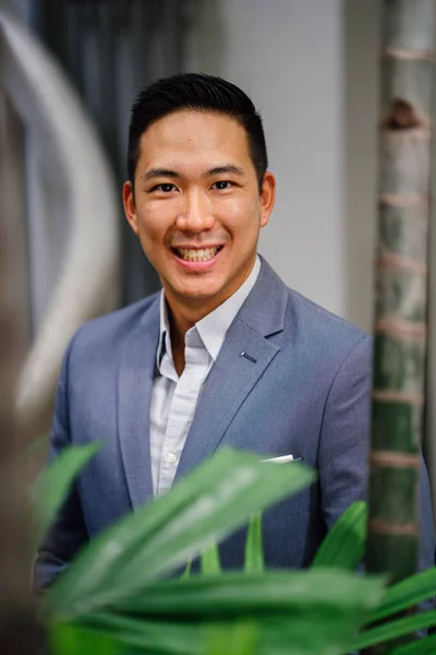 Portrait of a handsome, young, smart Chinese Asian man in a suit smiling and standing against a green wall in the day. He has short hair and is wearing a grey suit.