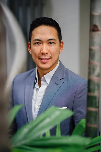 Portrait of a handsome, young, smart Chinese Asian man in a suit smiling and standing against a green wall in the day. He has short hair and is wearing a grey suit.