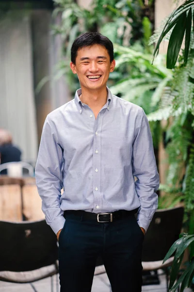 Portrait of a handsome, young, smart Chinese Asian man in a suit smiling and standing against a green wall in the day. He has short hair and is wearing a grey suit.