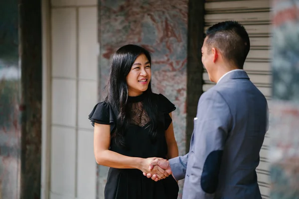 Two Chinese business people (a man in a suit and professionally dressed woman) shake hands in agreement and accord over a deal in the daytime against a plain background.