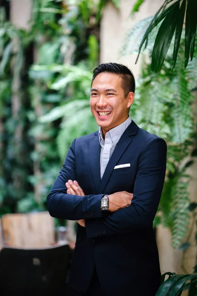 Portrait of a handsome, young, smart Chinese Asian man in a suit smiling and standing against a green wall in the day. He has short hair and is wearing a grey suit.