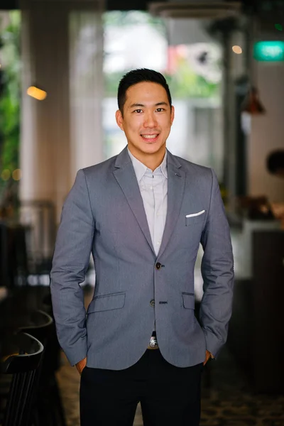 Portrait of a young, professional and handsome Chinese Asian lawyer. He is in a grey suit and white pocket square and is smiling at the camera. He is well groomed and put together and is smiling.