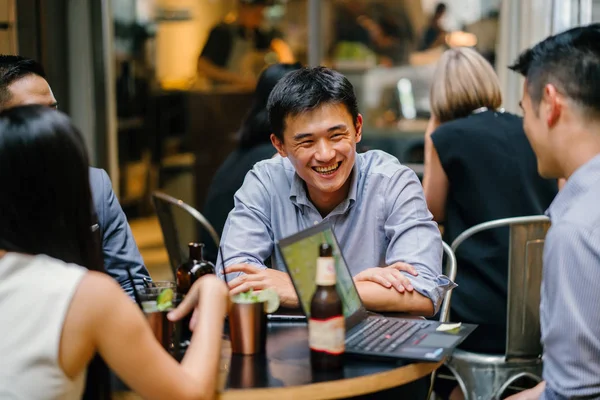 group of young and energetic Chinese Asian coworkers sit around a table and have a light hearted business discussion. They are smiling and laughing as they talk and have a conversation.