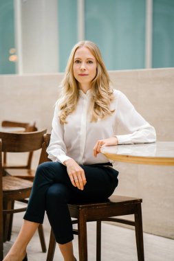 Doğal ışık dolu bir ofiste gün boyunca bir masada oturan bir genç, beyaz Avrupa kadın portresi. O sarışın, mavi gözlü, var ve profesyonel olarak giyinmiş ve gülümsüyor