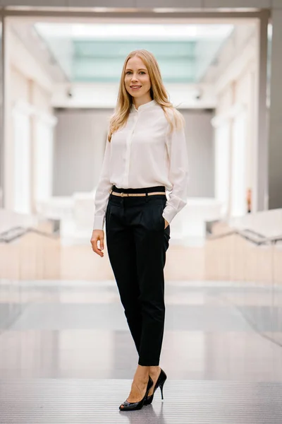 full-length, full-body shot of an attractive mature Russian woman leader. She is elegantly dressed in professional white shirt and black pants and heels and is smiling confidently.