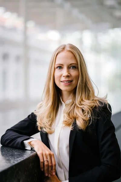 Russian woman in a suit leans against a stone in the day and is smiling. She is blond with blue eyes and is attractive and professional in a black suit and white shirt.