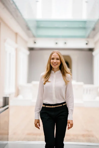 full-length, full-body shot of an attractive mature Russian woman leader. She is elegantly dressed in professional white shirt and black pants and heels and is smiling confidently.