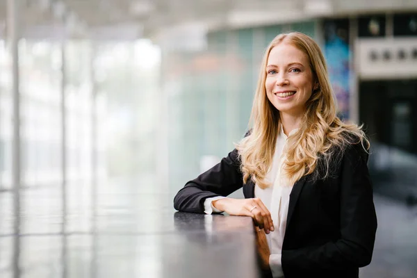 Russian woman in a suit leans against a stone in the day and is smiling. She is blond with blue eyes and is attractive and professional in a black suit and white shirt.