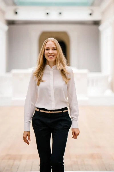 full-length, full-body shot of an attractive mature Russian woman leader. She is elegantly dressed in professional white shirt and black pants and heels and is smiling confidently.