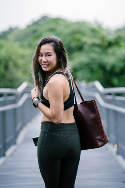 Portrait of an attractive, young Chinese Asian woman dressed in sporty, active wear with bag in green public park in Singapore during the day on the weekend.
