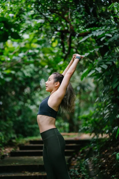 young Chinese Asian stretching before her weekend workout. She is in exercise gear and is in a lush, green forest in Asia during the day.