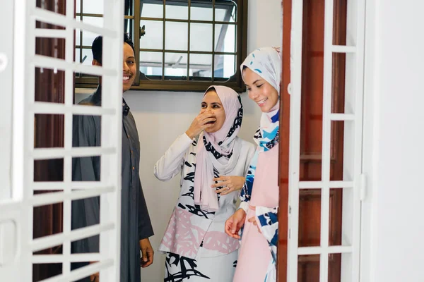 Muslim husband and wife couple greet a guest at their door and invite her in to celebrate Hari Raya, a Muslim celebration of love and forgiveness. They are overjoyed to see one another.