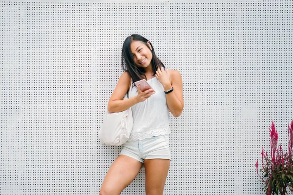 candid portrait of a cute, athletic and tanned Chinese Asian woman using her smartphone in the day on a street. She is texting and messaging her friend while she waits for someone.