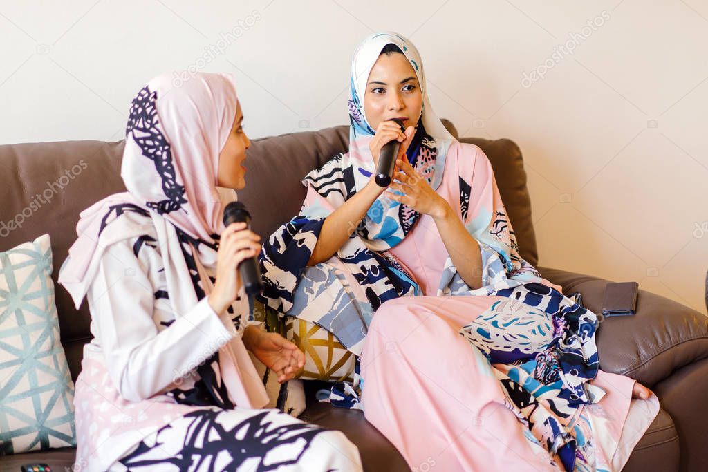 Two Muslim women in headscarves are singing karaoke on their home entertainments system over Hari Raya and enjoy each other's company.