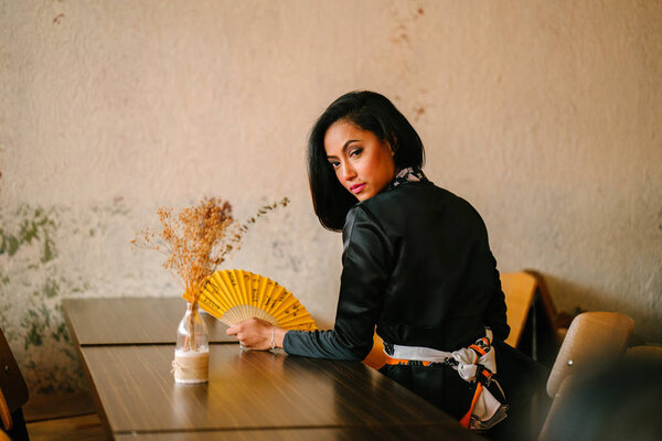 Portrait of a stylish, fashionable and slim Malay Muslim woman in a cafe or coworking space during the day. She is holding a fan and sitting at a desk.