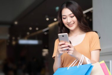 Alışveriş merkezinde alışveriş yaparken akıllı telefon kullanan mutlu kadın alışverişi; dijital cüzdan teknolojisi, elektronik ödeme, akıllı telefon siparişi, modern alışveriş teknolojisi kavramı