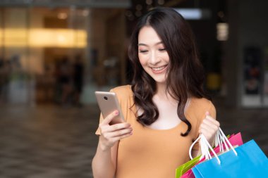 Alışveriş merkezinde alışveriş yaparken akıllı telefon kullanan mutlu kadın alışverişi; dijital cüzdan teknolojisi, elektronik ödeme, akıllı telefon siparişi, modern alışveriş teknolojisi kavramı