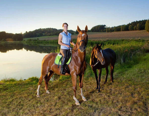 Женщина с двумя лошадьми у озера на закате — стоковое фото