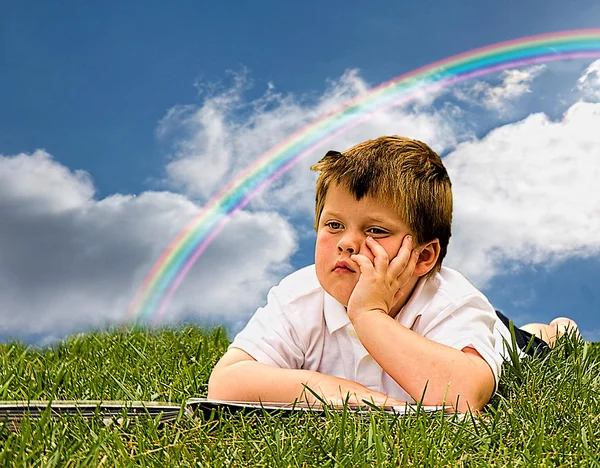 Kleine jongen met boek terwijl dromen op gras en regenboog in de rug — Stockfoto