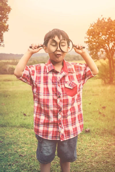 Asian boy with magnifying glass at park on vacation. Warm tone. — Stock Photo, Image