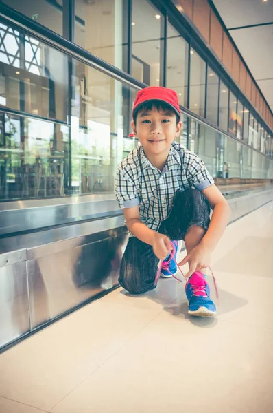 Happy asian child sitting and tying to tie shoelaces near walkway at airport. — Stock Photo, Image