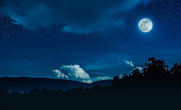 Landscape of blue night sky with many stars and cloudy above silhouette of mountain range and trees. Beautiful bright full moon over tranquil nature on dark tone. The moon taken with my own camera.