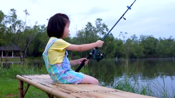 穏やかな夜のラグーンのほとりに座っている愛らしいアジアの子供 少女は釣竿を持っている — ストック動画